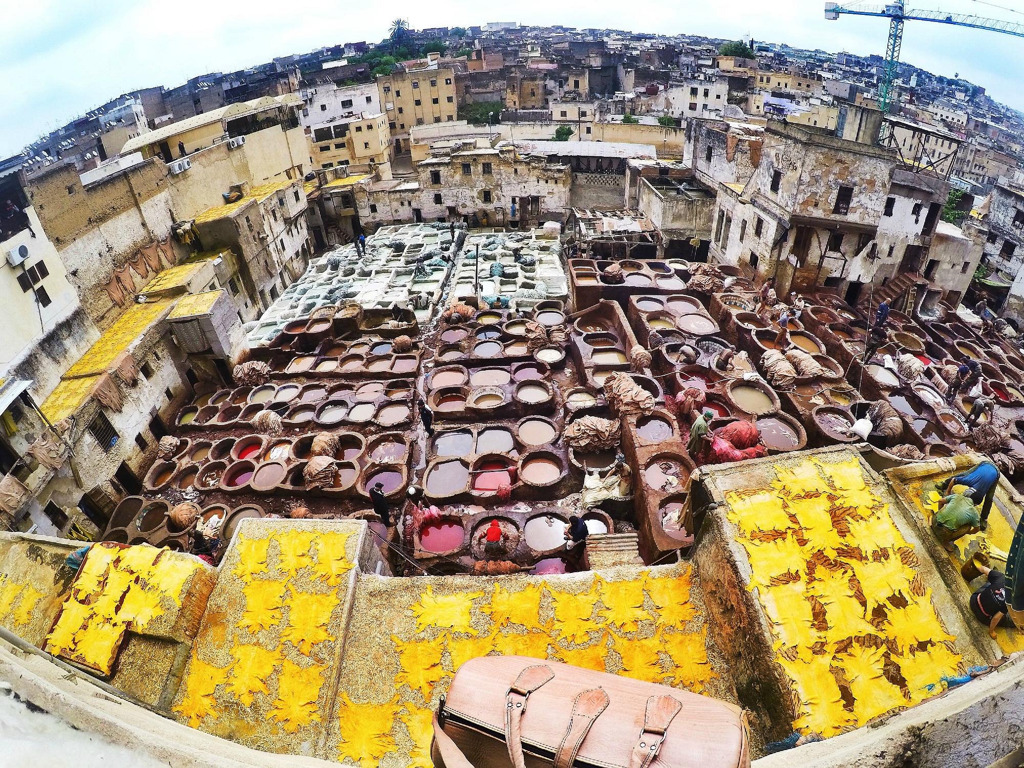 Aerial view of Chouara Tannery in Fez, Morocco, showing rows of colorful dye vats, workers handling leather, and yellow hides drying on rooftops.