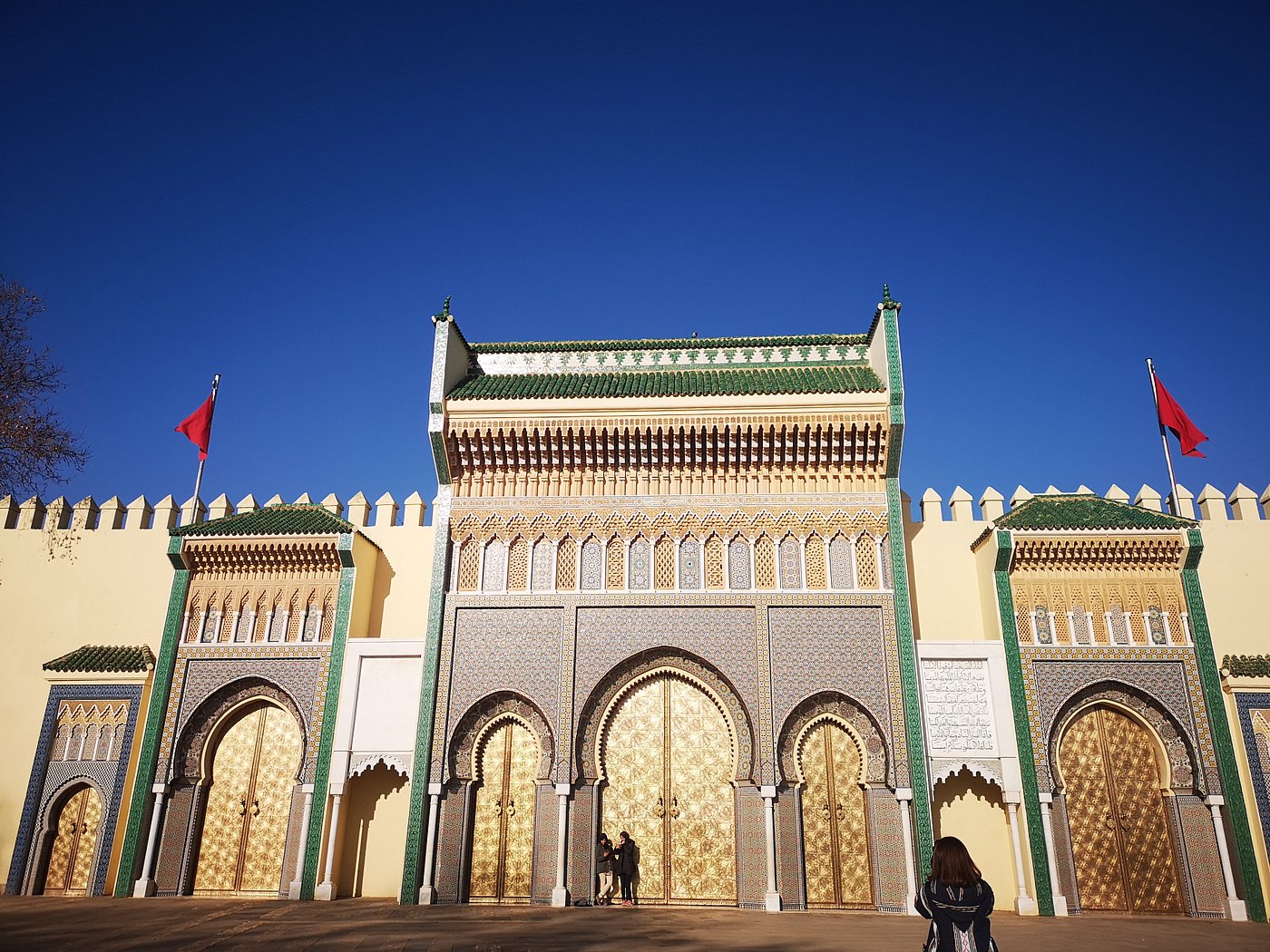 olden Gates of the Royal Palace – Fez, Morocco Front view of the ornate golden gates of the Royal Palace in Fez, Morocco, with intricate mosaic tilework, green rooftops, and two Moroccan flags under a clear blue sky.