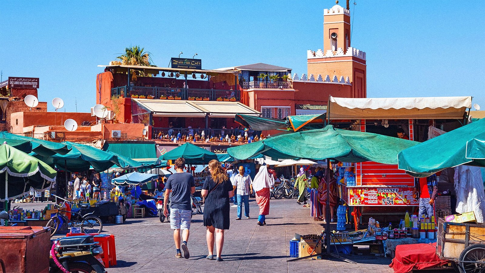 Two tourists walking through Jemaa El Fna square in Marrakech, surrounded by colorful market stalls and local vendors.