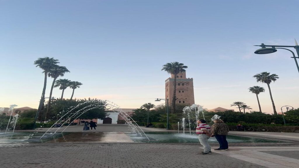 Sunset view of the Koutoubia Mosque in Marrakech with palm trees and fountain jets in the foreground.